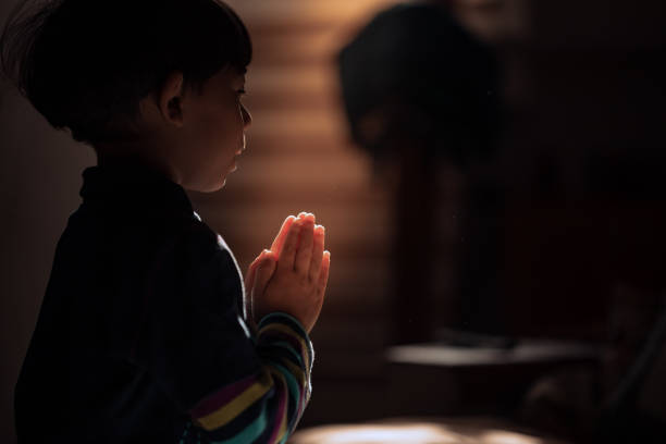 Child on a dark background praying to God, a ray of light illuminates the joined hands. Concept of religion and faith
