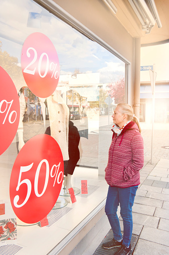 Woman with long blond hair in casual clothing looks into a shop window of a fashion store with discount signs and is thinking about to buy a new coat, which is wishful thinking.