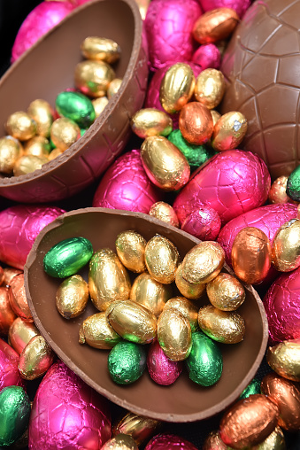 Pile or group of multi colored and different sizes of colourful foil wrapped chocolate easter eggs in pink, red, gold and lime green with two halves of a large brown milk chocolate egg in the middle and mini eggs inside.