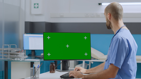 Physician assistant sitting at desk working at sickness expertise typing medical report on mock up green screen chroma key computer with isolated display in hospital office. Medicine concept