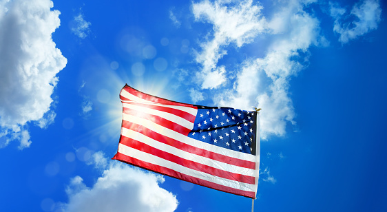 Conceptual image of waving American flag at flagpole over sunny blue sky with clouds and shinning sun