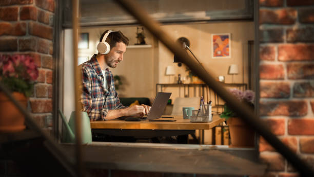 istock Handsome Caucasian Man Working on Laptop Computer, Sitting Behind His Desk at Home Office. Freelance Entrepreneur Listening to Music, Doing Remote Telework. View From Outdoors into Window. 1461320687