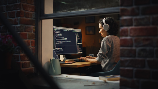 Hispanic Female Programmer in Headphones Listening Podcast and Working on Desktop Computer, Screen Shows Coding Language User Interface. Digital Entrepreneur Creating Modern Software.