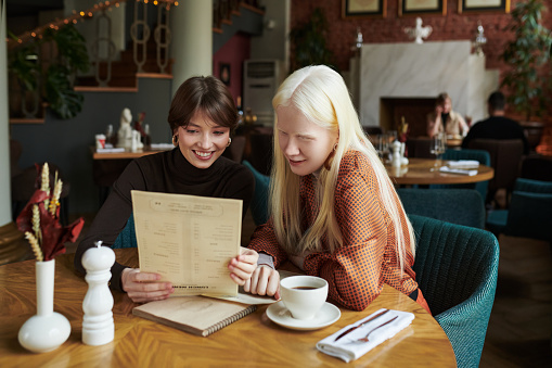 Albino girl and her happy friend looking through menu and choosing food while sitting by table in cozy cafe during their meeting at leisure