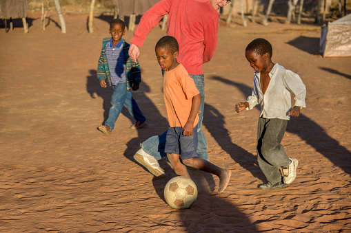 African village children playing football on the dirt in the yard with a Caucasian young adult