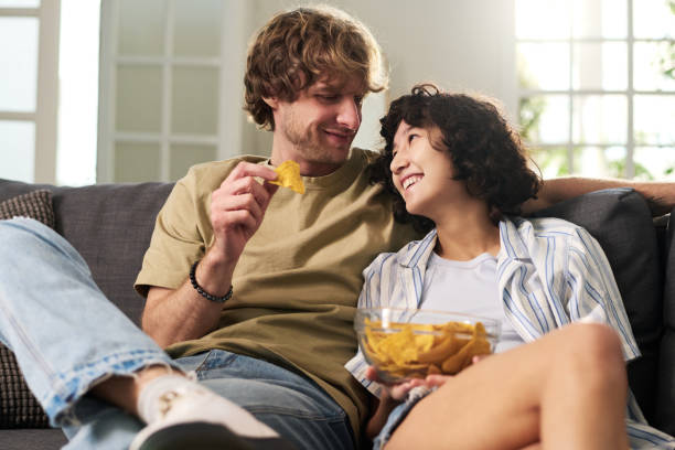 Young affectionate man and woman looking at one another Young affectionate man and woman looking at one another while relaxig on comfortable sofa in living room at leisure and having potato chips family eating potato chips stock pictures, royalty-free photos & images