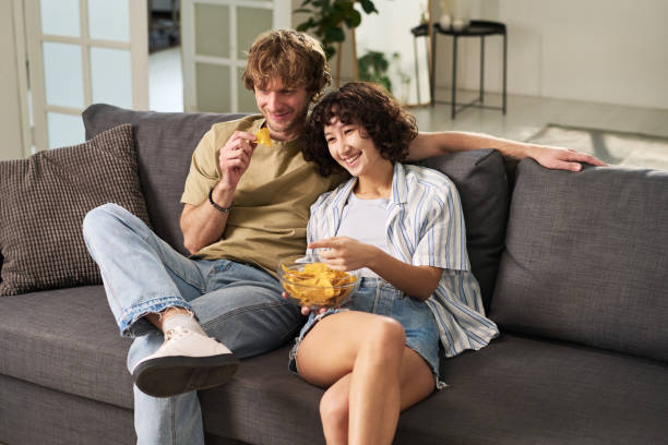 Happy young husband and wife laughing while watching comedy Happy young husband and wife laughing while sitting on couch in front of tv set, eating potato chips and watching comedy at leisure family eating potato chips stock pictures, royalty-free photos & images