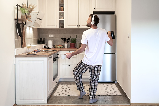 Bearded man in pajama pants and white t-shirt dances with headphones in domestic kitchen in the morning while making coffee.