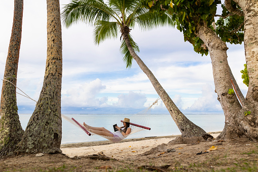 A shot of a woman sat in a hammock on a tropical beach reading a book. It is a bright sunny day and she is sat in the hammock under two palm trees wearing a sunhat.  The beach is located in Fiji.