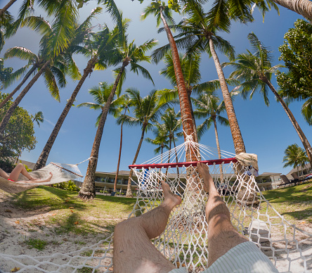 A waist down shot of two people laying in hammocks underneath palm trees on a tropical holiday resort in Fiji. The camera is pointing up into the sky. It is a bright sunny day.