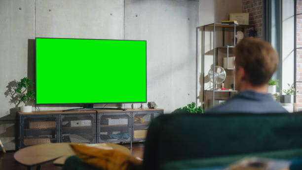 istock Young Attractive Caucasian Man Sitting on a Sofa and Watching TV with Horizontal Green Screen Mock Up. It's Day Time on Weekend at Home. Spacious Living Room in Stylish Loft Apartment. 1461341617