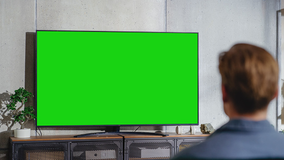 Young Attractive Caucasian Man Sitting on a Sofa and Watching TV with Horizontal Green Screen Mockup. It's Day Time on Weekend at Home. Spacious Living Room in Stylish Loft Apartment.