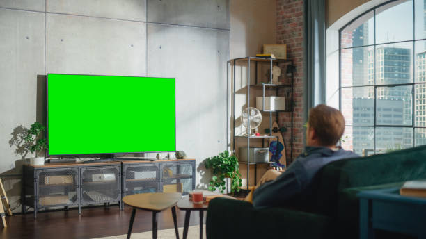 istock Young Handsome Caucasian Man Sitting on a Sofa and Watching TV with Horizontal Green Screen Mock Up. It's Day Time on Weekend at Home. Spacious Living Room in Stylish Loft Apartment. 1461341662