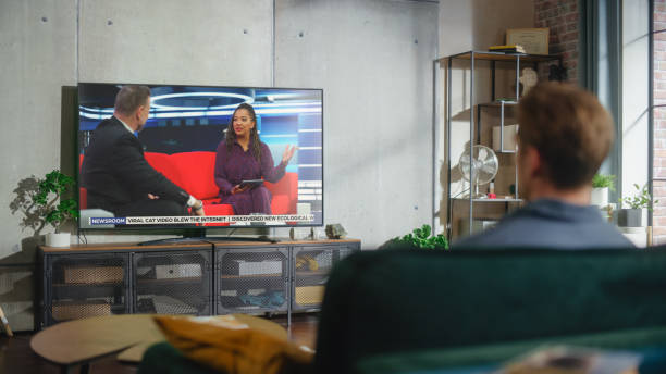 istock Young Handsome Man Sitting in Relaxed Pose on a Sofa and Watching TV with Live Talk Show During the Day Time on Weekend. Serious Female African American Host Interviews Guest in Studio. 1461341713