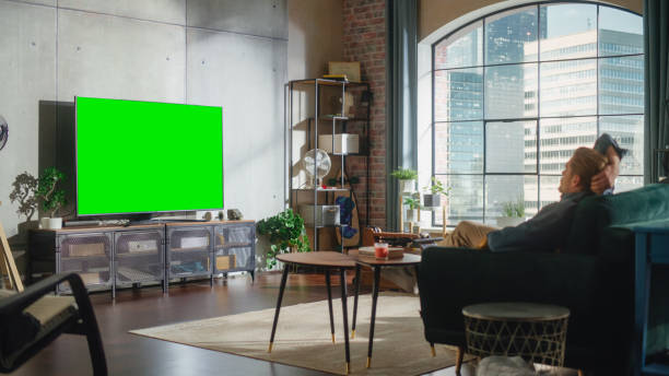 istock Young Handsome Man Sitting on a Sofa and Watching TV with Horizontal Green Screen Mock Up. It's Day Time on Weekend at Home. Spacious Living Room in Stylish Loft Apartment. 1461341766