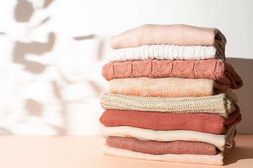 Neat stack of textured woolen sweaters in pastel beige, pink and orange colors against sunlit white background. Autumn and winter fashion aesthetic.