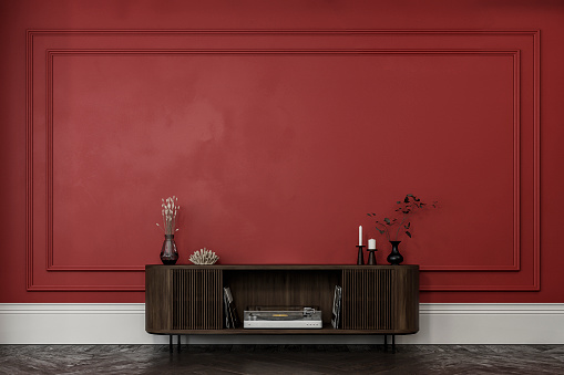 Empty classic elegant interior with a french style antique red wall background with moldings (frame), decoration on a low hardwood cabinet (gramophone, gramophone records, vase, candle holder, dry flowers) on the dark brown herringbone hardwood floor, and copy space. 3D rendered image.