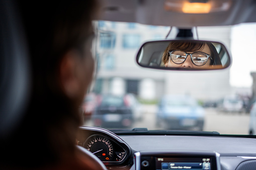 Adult caucasian businesswoman in glasses looking in the rearview mirror while sitting at the wheel of a car