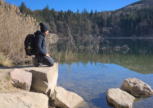 Middle-aged man backpacking alone in a wool jumper and hat sits on his knees in front of a mountain lake in winter