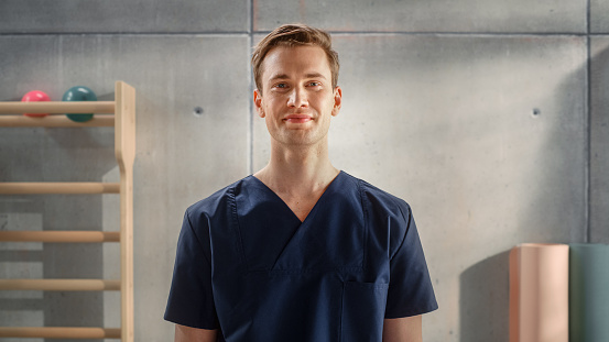 Portrait of a Handsome Professional Male Nurse in Uniform Standing and Posing for Camera, Smiling in Medical Rehabilitation Center. Physiotherapy Assistant In Hospital. Young Practising Doctor.