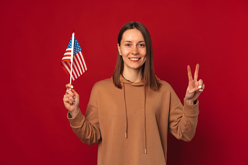 Cheerful young woman is holding up USA flag and two fingers peace gesture over red background.