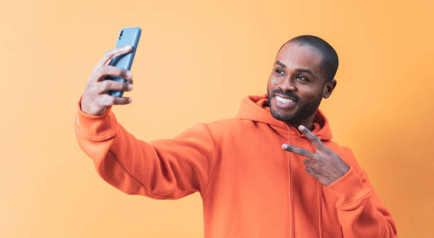 istock A handsome young Colombian man takes a selfie with his mobile while doing the victory gesture and smiling, the man is wearing an orange hoodie. 1461354872