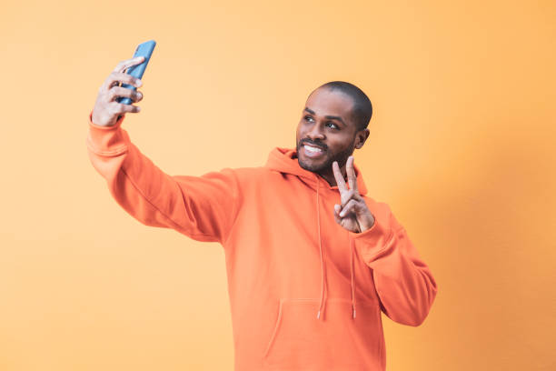 istock A handsome young Colombian man takes a selfie with his mobile while doing the victory gesture, the man is wearing an orange hoodie, orange background. 1461354875