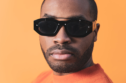 Portrait of a serious Colombian young man, wearing an orange t-shirt and sunglasses. Black man isolated on orange background. Male accessories.