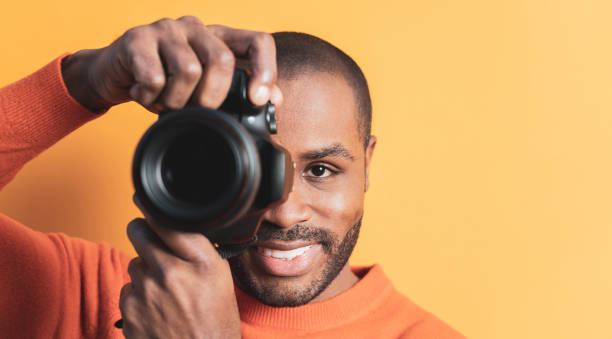 istock A portrait of a smiling dark-skinned young man posing with a digital camera, orange colored background. photographer,copyspace. 1461354983