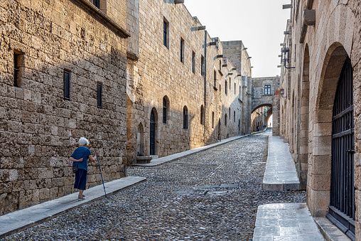 Rhodes Island, Greece - May 25, 2022: An old woman with white hair a cane walking uphill on the street of the Knights in the historic old town of Rhodes, Greece. The small stones laid on the road were made so that the feet of the horses would not slip.