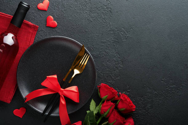 istock Valentines day romantic table setting. Empty closeup of red roses, wine, candles, dinner black plate, knife, fork and decorative silk hearts on black background. Holiday concept. Copy space. Top view 1461356947