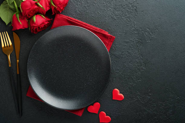 istock Valentines day romantic table setting. Empty closeup of red roses, wine, candles, dinner black plate, knife, fork and decorative silk hearts on black background. Holiday concept. Copy space. Top view 1461356973