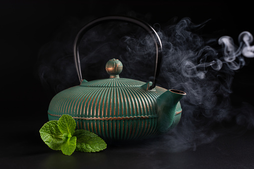 Traditional green asian cast iron hot teapot or kettle with steam and mint leaves on dark background