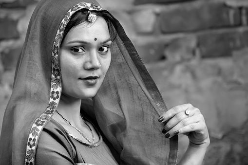 Black and white close-up portrait of a beautiful, traditional rural Indian woman in north Indian attire and covering her head with dupatta. She is standing against a concrete wall and looking at the camera with a smile.