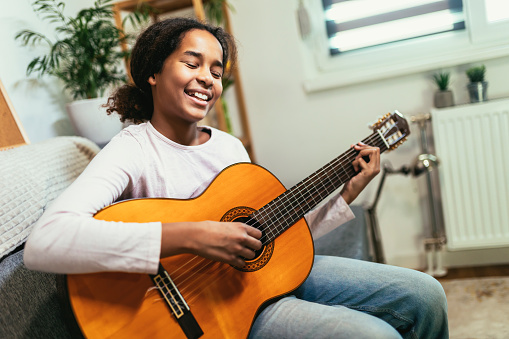 Smiling multi-ethnic females playing ukulele in class. Instructor teaching music to pre-adolescent student. They are sitting in classroom at education building.