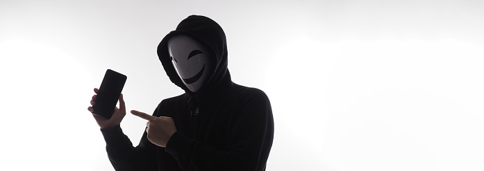 Hacker Anonymous and face mask with smartphone in hand. Man in black hood shirt holding and using mobile phone on white background. Represent cyber crime data hacking or stealing personal data concept