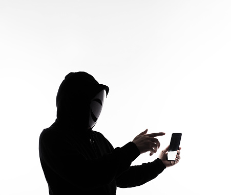 Hacker Anonymous and face mask with smartphone in hand. Man in black hood shirt holding and using mobile phone on white background. Represent cyber crime data hacking or stealing personal data concept