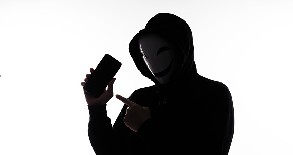 Hacker Anonymous and face mask with smartphone in hand. Man in black hood shirt holding and using mobile phone on white background. Represent cyber crime data hacking or stealing personal data concept