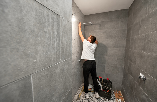 Male plumber fixing bathroom showerhead while working on bathroom renovation. Back view of man worker installing high pressure bath showerhead at home.
