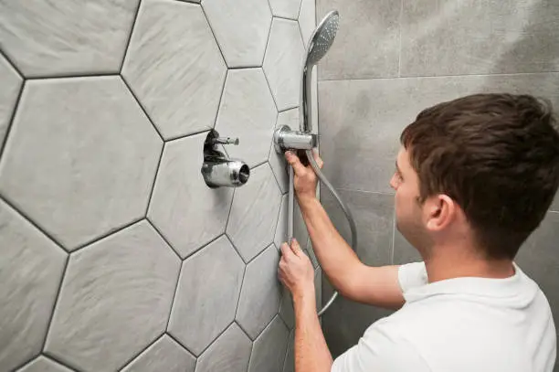 Male worker fixing metal bath showerhead and doing plumbing works while working on home renovation. Man standing by the wall with ceramic tile and installing handheld showerhead in bathroom.