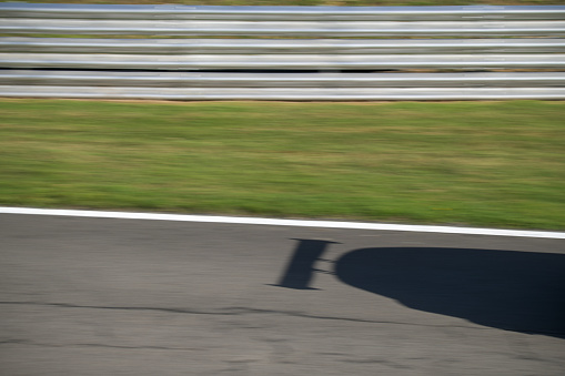 The shadow of the rear of a race car and it’s spoiler passing the silver barrier at speed on a tarmac race circuit