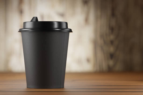 A black disposable machine with a plastic lid on a wooden table. Mockup of a black disposable glass. Black paper cup mockup for your design. 3d render. stock photo