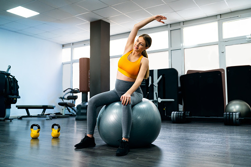 Young pregnant Caucasian woman, sitting on fitness ball and stretching her body in the gym