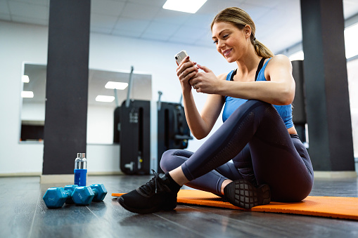 Young pregnant Caucasian woman, sitting on yoga mat and using mobile phone in the gym