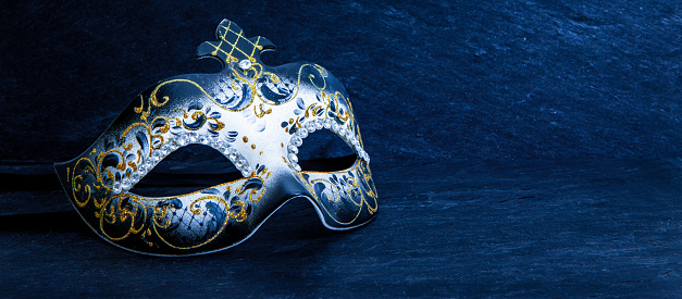 Front side view of black and white Venetian mask decorated with gold glitter decorations and diamond beads around the eye area. Masquerade and carnival concept. Festive mask isolated on dark blue slate stone background.