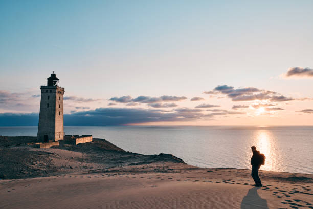 Silhouette man standing on sandy beach with scenic view of seascape and lighthouse on coast against sky during sunset