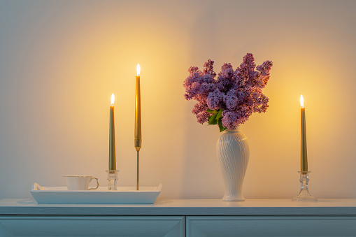 lilac flowers in white vase and golden candles on background white wall