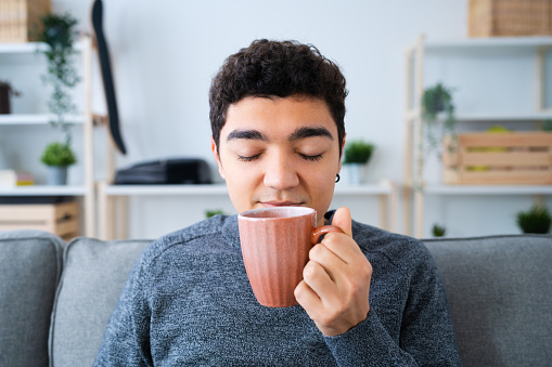 Hispanic teenager boy holding cup of hot drink and smelling it in the living room