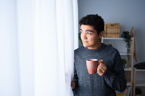 Portrait of confident hispanic teenager boy looking through the window and holding cup of coffee