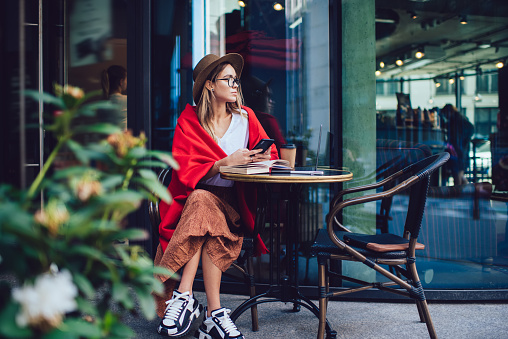 Thoughtful woman in hat and glasses warming with colourful red blanket and surfing smartphone while sitting by table with opened book and coffee cup at street cafe